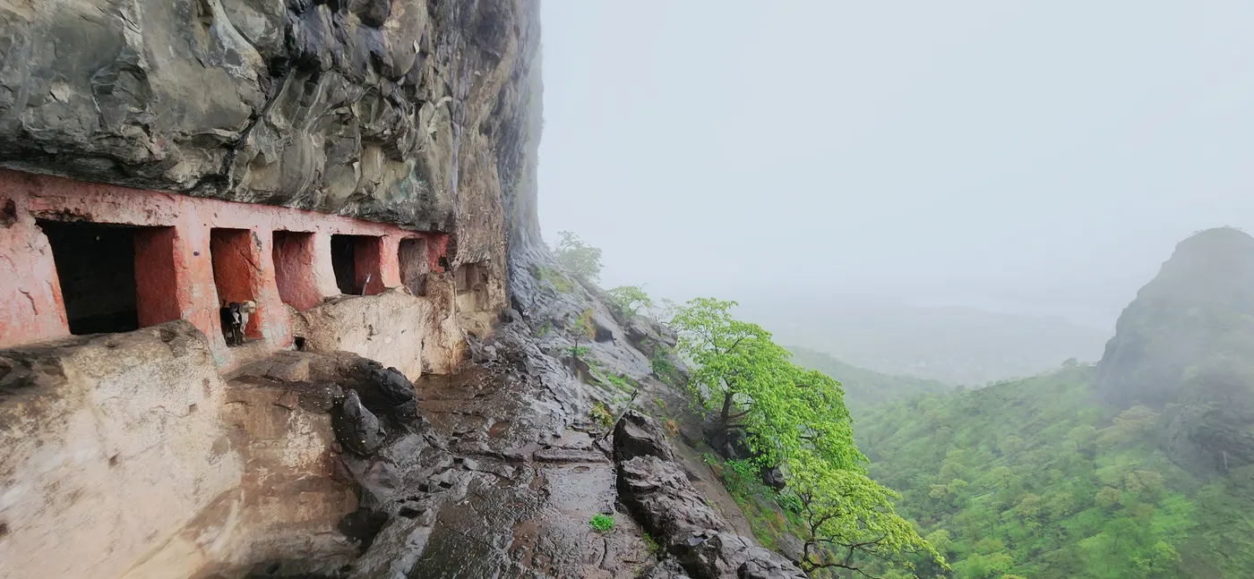 Rock-cut cave openings with red pillars carved into a wet cliff, the valley falling away into mist beside them.