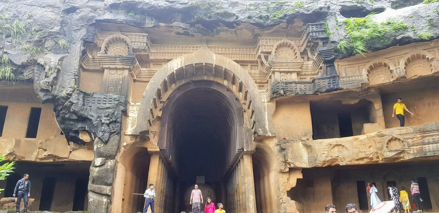 The carved stone front of a cave, with a tall arched doorway and ornate rock carvings, and visitors standing in front.
