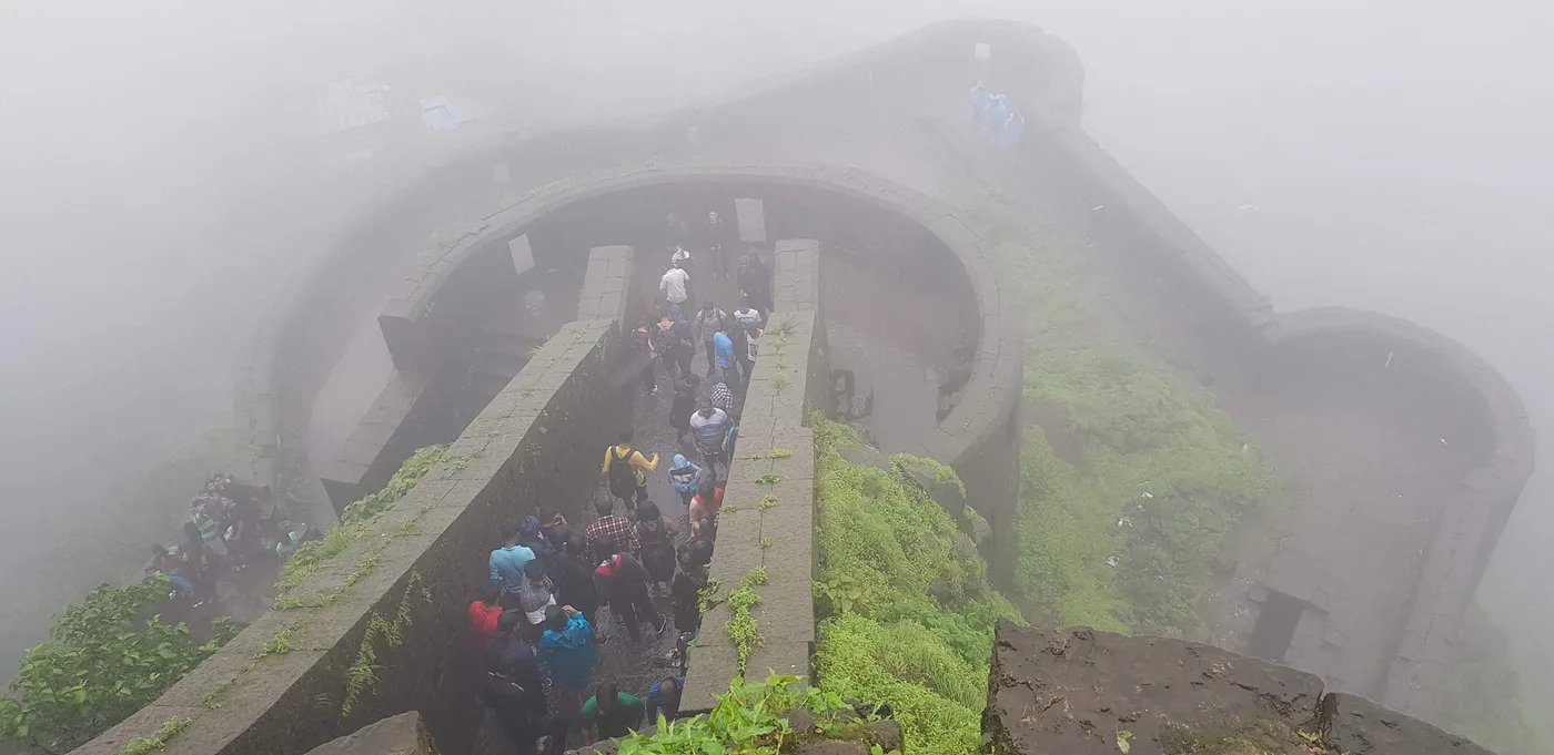 Looking down from a fort wall in thick fog at a crowd climbing through a curved stone gateway.