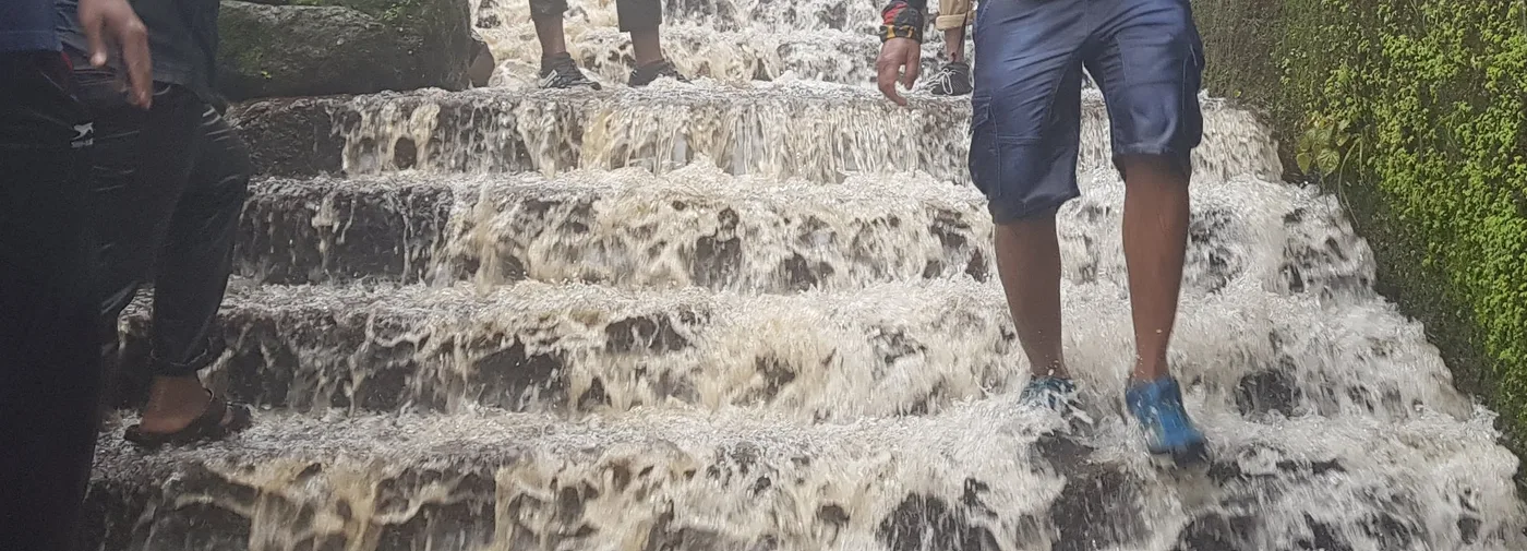 Water pouring down a flight of stone steps beside a mossy green wall, with someone walking through it.